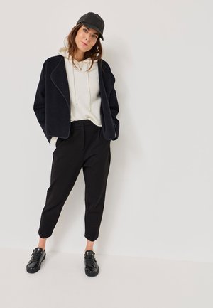 Navy oversized jacket, white hoodie with drawstring, black tapered trousers, and shiny black sneakers. Model wears a black cap.