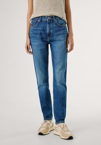 Blaue Denim-Jeans mit schlanker Passform, fünf Taschen und Knopfverschluss. Kombiniert mit beigen Sneakers und einem strukturierten grauen Oberteil.