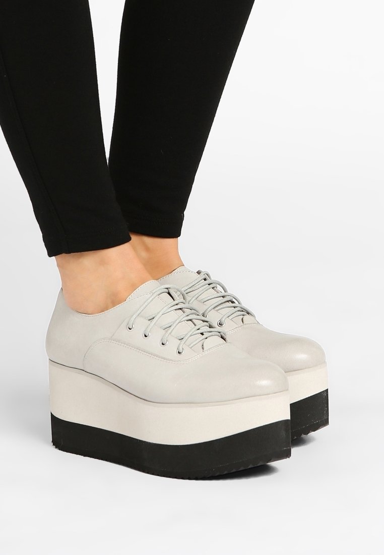 Pieds chaussés de chaussures beiges à plateforme à lacets avec semelles épaisses noires et blanches, assorties à un pantalon noir ajusté.