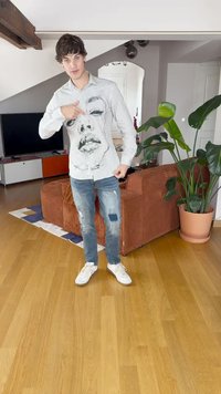 Λευκό πουκάμισο μακρυμάνικο με μαύρη εκτύπωση προσώπου, συνδυασμένο με φθαρμένα μπλε τζιν και λευκά αθλητικά παπούτσια. Το φόντο περιλαμβάνει εσωτερικά φυτά και έπιπλα.