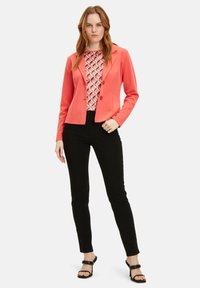 Korallenfarbener Blazer mit zwei Knöpfen, kombiniert mit einem gemusterten Oberteil mit geometrischen Formen in Rosa und Orange. Schwarze eng geschnittene Hosen und hohe Schuhe.