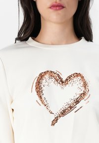Kremfarget genser med et hjerte-design laget av paljetter i nyanser av roségull og sølv, med et glatt teksturert stoff.