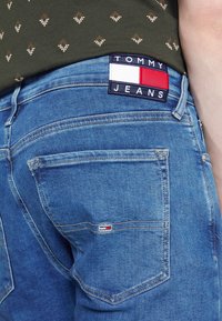 Modré džíny z denimu s červenou, bílou a námořnicky modrou etiketu s nápisem "TOMMY JEANS" na pase a malým logem na zadní kapse.