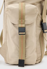 Beige rugzak gemaakt van nylon met een grote voorzak. Heeft dikke, gestructureerde banden met rode stiksels en een zwarte kunststof gesp.