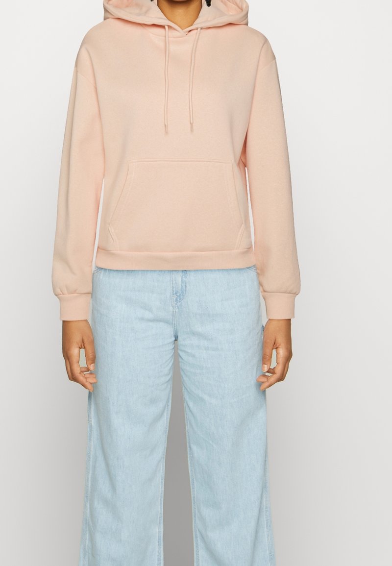 Lichtroze hoodie met een voorzak, geribbelde manchetten en een ontspannen pasvorm, gecombineerd met lichtblauwe wijduitlopende jeans.