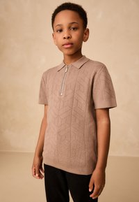 Tricou polo cu mâneci scurte și fermoar, din material tricotat bej cu model geometric. Guler simplu și detalii texturate pe întreaga suprafață a corpului.