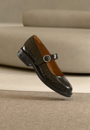 Chaussure Mary Jane en cuir noir perforé avec une boucle ronde en argent, présentée en suspension au-dessus d'un tapis beige et d'un canapé en arrière-plan.