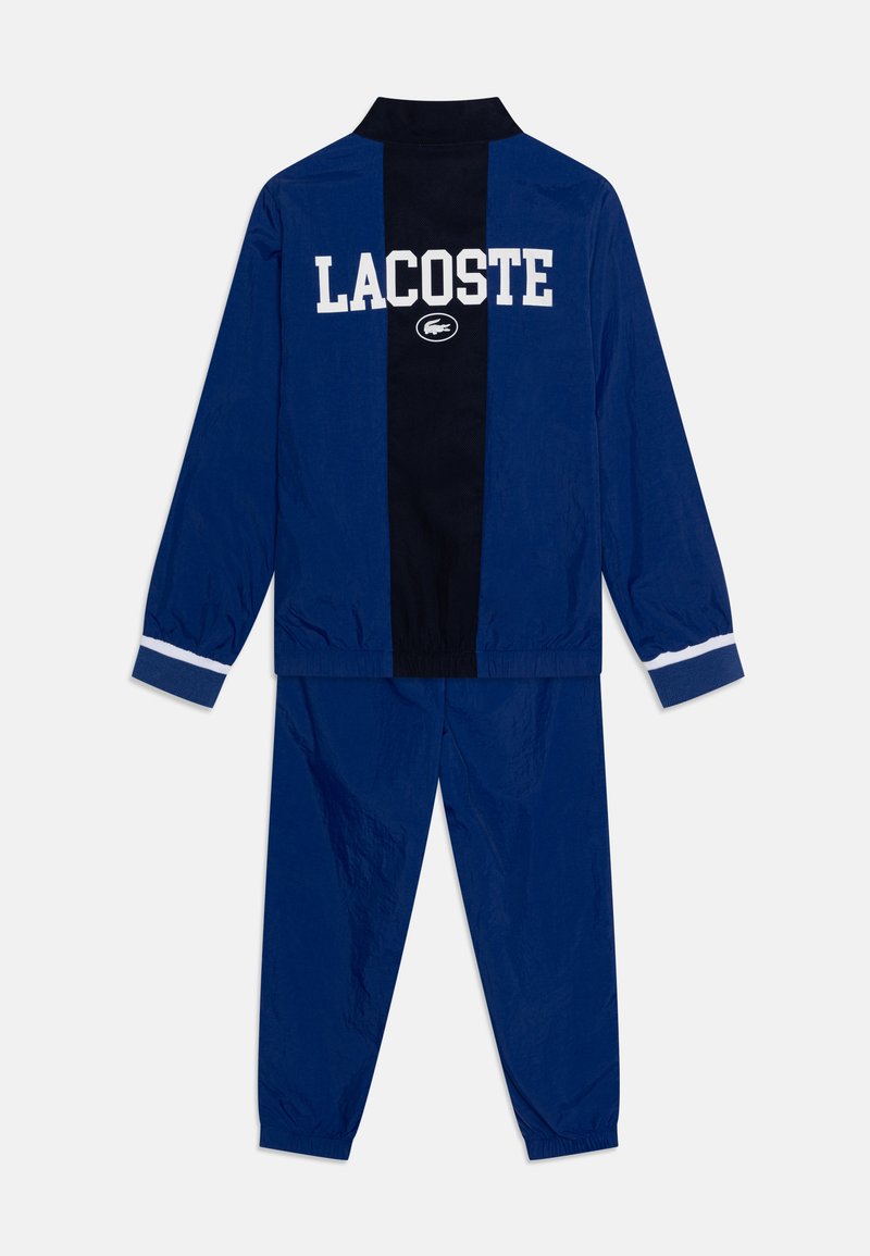 Survêtement marine comprenant une veste zippée avec le texte "LACOSTE", un écusson de logo, et un pantalon assorti avec des poignets et une taille élastiques.