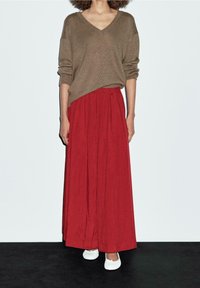 Ruskea neule v-pääntiellä yhdistettynä punaisiin, laskostettuihin maksihameeseen. Mallilla on valkoiset litteät kengät. Tausta on tasaisen valkoinen.