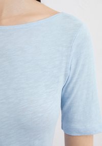 Světle modrý top s krátkým rukávem, vyrobený z měkké, texturované látky; má široký, zaoblený výstřih a mírně volný střih.