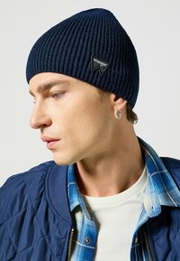 Beanie a coste navy con una toppa triangolare nera "Wrangler", indossata da un modello che porta una giacca blu trapuntata e una camicia a quadri.