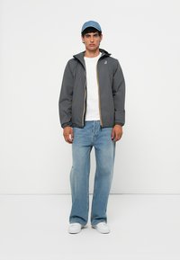 Chaqueta ligera gris con cremallera y ribete colorido, usada sobre una camiseta blanca. Jeans de mezclilla azules y zapatillas blancas completan el atuendo.