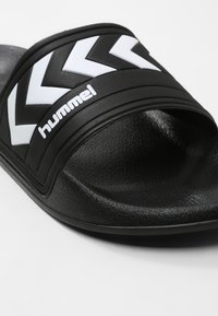Svart halksandaler med en texturerad sula och en tjock rem med vita chevronmönster och "hummel"-logotypen.