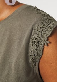 Ærmeløs olivengrøn top med indviklet blondering på skuldrene, båret af en person med medium-mørk hud og runde hvide øreringe.