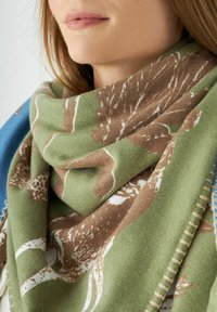 Écharpe à motifs verts et bruns avec une texture douce, présentant des motifs inspirés de la nature et des accents blancs, drapée autour du cou.