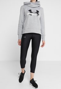 Vrouw in een grijze Under Armour hoodie, zwarte fitted leggings en zwarte sportieve schoenen, staand tegen een eenvoudig witte achtergrond.