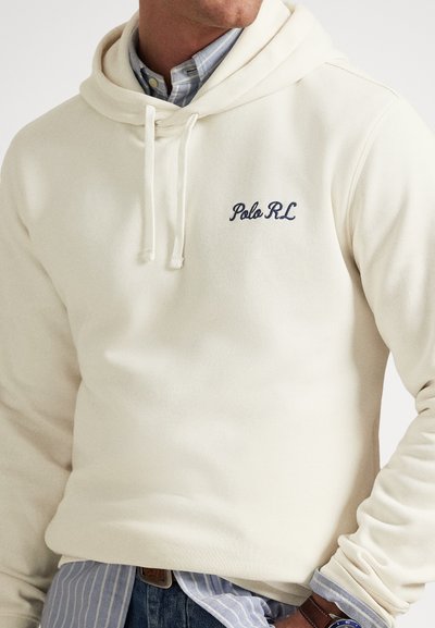 Muž nosí krémovú mikinu Polo RL s kapucňou cez modrú pruhovanú košeľu na gombíky a rifle, na zápästí má viditeľné hnedé kožené hodinky.
