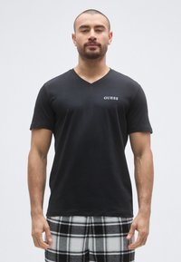 Sort t-shirt med v-udskæring og "GUESS"-logo på brystet, lavet af blødt materiale. Modellen kombinerer den med sorte og hvide ternede pyjamasbukser.
