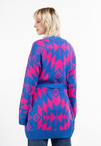 Dlhý modrý kardigán s výraznými ružovými geometrickými vzormi. Má opasok v oblasti pásu a rebrované manžety. Mäkká pletená textúra.