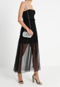 Robe noire bustier avec un voile transparent, dotée d'un corsage texturé et d'une taille à volants. Associée à des sandales vertes à talons hauts et une pochette argentée.