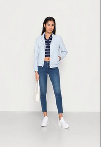 Casaco bomber azul-claro, top crop riscado em azul-marinho, jeans azuis justos com painéis, sapatilhas brancas e uma pequena bolsa branca.