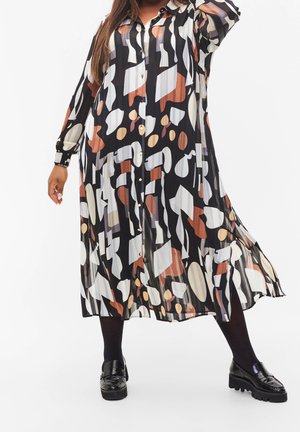 Personne portant une longue robe noire plissée avec des formes abstraites beige, blanches et brunes, des collants noirs et des mocassins noirs épais.