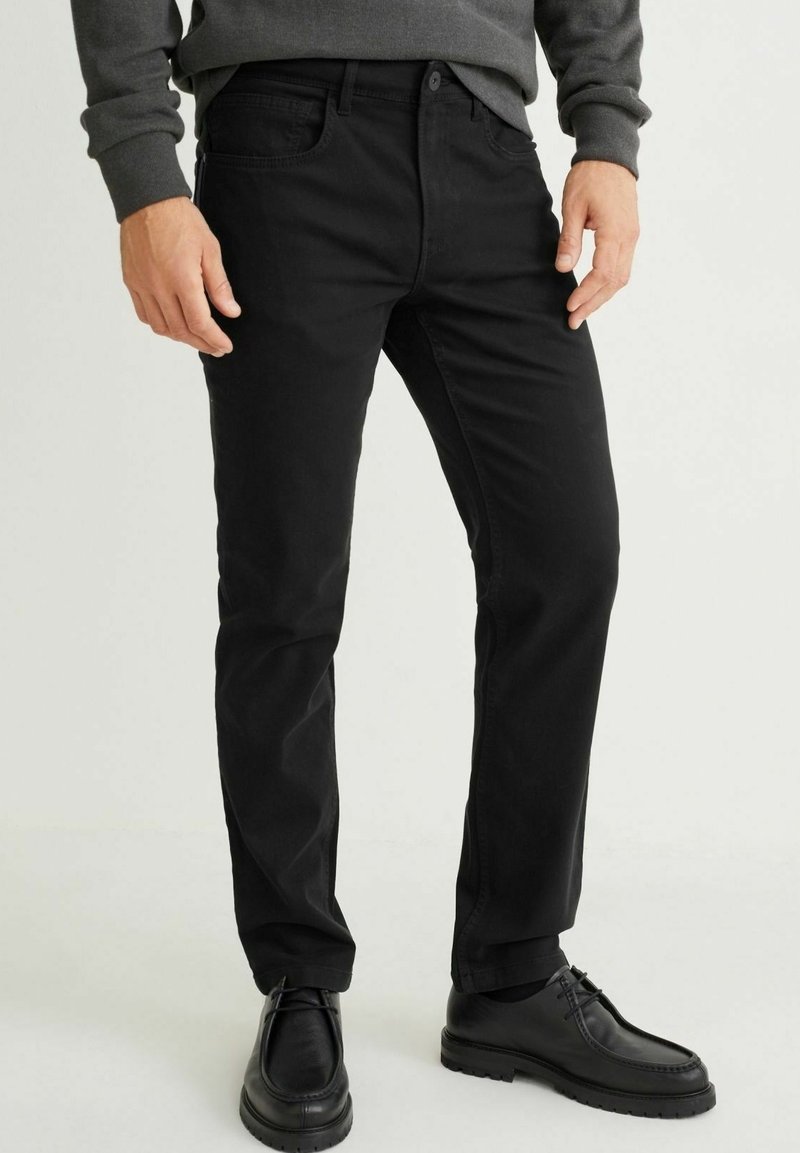 Pantalon en denim noir à coupe slim, design à cinq poches, fermeture par boutons. Porté avec des chaussures noires, sur un fond neutre.