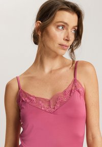 Vaaleanpunainen toppi, jossa on säädettävät olkaimet ja pitsiyksityiskohta kaula-aukossa. Kangas on sileä ja siinä on hieno kiilto, mikä luo herkän ilmeen.