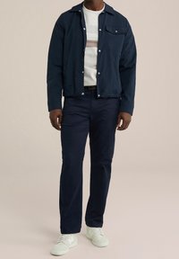 Veste légère bleu marine avec une poche avant, portée sur une chemise gris clair à rayures horizontales, associée à un pantalon bleu marine et des baskets blanches.
