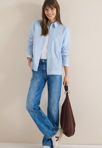 Femme souriante, portant une chemise boutonnée rayée bleu et blanc, un haut blanc, un jean bleu, des baskets blanches, tenant un sac en daim marron.