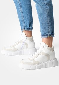 Baskets montantes blanches faites de cuir lisse et de daim, avec une semelle épaisse et rainurée, des accents beiges contrastants et une fermeture éclair sur le côté.