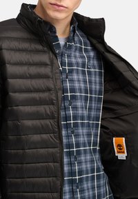 Giacca nera imbottita con colletto alto, indossata aperta sopra una camicia a quadri blu e bianca. Include un'etichetta arancione Timberland all'interno.