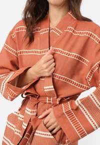 Terakotový plášť s bielymi pruhovanými vzormi; vyrobený z textúrovanej látky, s širokými rukávmi a previazaným pásom pre nastaviteľné prispôsobenie.