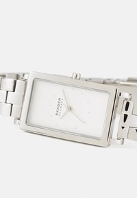 Silver rektangulär klocka med en vit urtavla, minimala timmarkörer och ett polerat metallarmband. Har en liten krona på sidan.