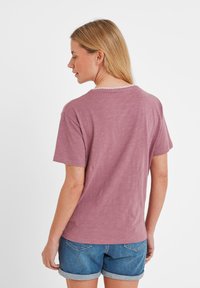 Woman with blonde hair wearing a short-sleeve mauve shirt and rolled-up denim shorts, facing away against a plain white background.