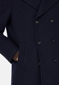 Marineblaue Woll-Peacoat mit einem doppelt-knöpfigen Design, schwarzen Knöpfen, großen Revers und einer Seitentasche. Strukturierter Stoff sichtbar.