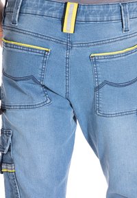 Pantalons cargo en denim bleu clair avec des accents jaunes. Parmi les caractéristiques, on trouve plusieurs poches, des coutures contrastées et une large ceinture avec une bande réfléchissante.