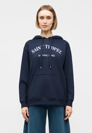 Nainen, joka pukeutuneena tummansiniseen huppariin, jossa teksti "Saint Tropez Michael Kors", ja leveälahkeiset siniset farkut, seisoo yksivärisen taustan edessä.