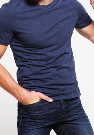 Mann trägt ein eng anliegendes marineblaues T-Shirt und dunkelblaue Jeans, mit einer Hand in der Tasche und der anderen erhoben.