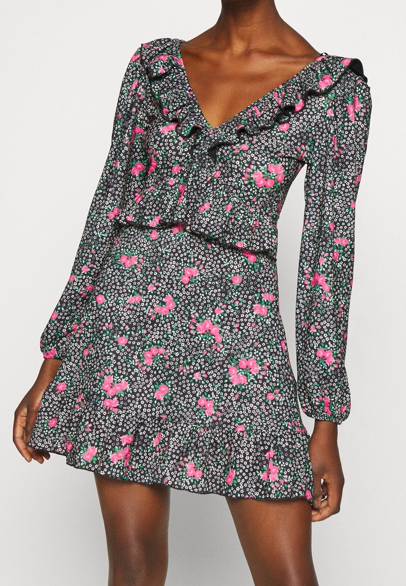 Mujer con un vestido negro de flores, con flores rosas y blancas, mangas largas, escote en V con volantes y una falda corta acampanada.