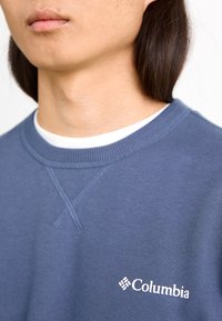 Granatowy sweatshirt z okrągłym dekoltem, ozdobiony detalami w postaci ściegów oraz białym logo Columbia na piersi.