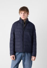 Jūros mėlynos spalvos pūkinė striukė su siuvinėtu dizainu, aukštu apykakle ir užtrauktuku. Yra subtili logotipo etikete ir kontrastingas užtrauktuko detalė.