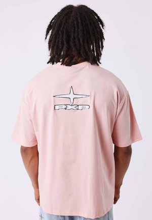 Personne avec des dreadlocks foncés portant un t-shirt rose clair avec une étoile blanche et le logo "PXP" dans le dos, debout devant un fond uni.