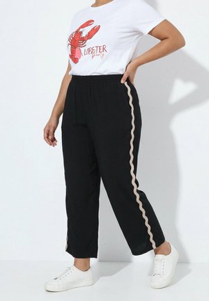 Person iført hvide sneakers, sorte bukser med beige bølgede sidestriber og en hvid t-shirt med en rød hummergrafik og teksten "Lobster Party".