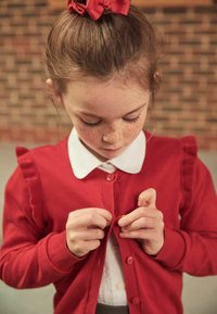 Red cardigan with ruffled sleeves, buttons, and a V-neck over a white collared shirt. The child is fastening the cardigan. Textured knit material.
