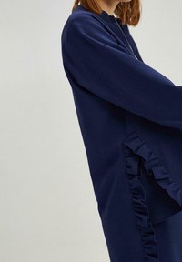 Marineblauwe sweatshirt met een rits aan de voorkant, voorzien van lange mouwen en een zijruffle-detail. Gemaakt van zacht materiaal met een gladde textuur.