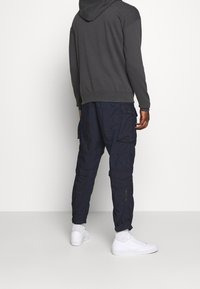 Sudadera gris con un bolsillo canguro, combinada con pantalones cargo azul marino que cuentan con múltiples bolsillos y un corte ajustado. Zapatillas blancas completan el conjunto.