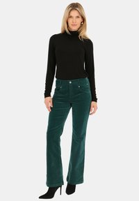 Zielone spodnie sztruksowe z wysokim stanem, kieszeniami z przodu i miękką fakturą, zestawione z czarną bluzką z długim rękawem oraz czarnymi wysokimi obcasami.