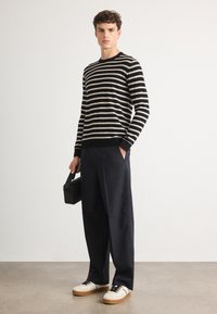 Suéter a rayas negro y crema, con corte relajado, combinado con pantalones de pierna ancha en azul marino. La modelo sostiene un bolso rectangular negro y lleva zapatillas blancas.