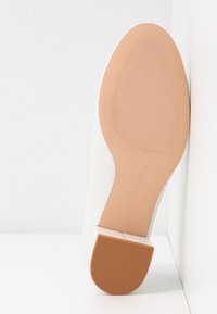 Vit högklackad sko med en slät läderfinish, fyrkantig tå och en ljust brun gummisåle med ett strukturerat greppmönster.